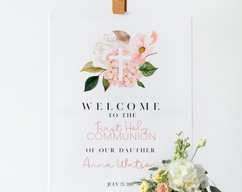 May include: A white sign with a floral design and a white cross. The sign reads "Welcome to the First Holy Communion of our daughter Anna Watson July 23, 2022".