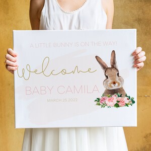 Puede incluir: Un cartel de lienzo blanco con texto rosa y dorado anunciando la llegada de "Baby Camila" el 25 de marzo de 2022. El cartel presenta una ilustración de acuarela de un conejo marrón con flores rosas.