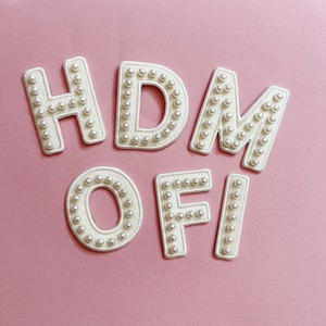 May include: HDM OFI spelled out in white felt letters with white pearl beads sewn around the edges on a pink background.
