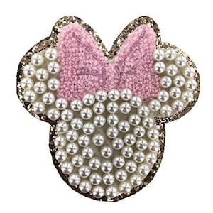 Pode incluir: Uma aplicação de tecido em forma de cabeça de rato de desenho animado com um laço rosa, adornada com glitter dourado e pérolas.