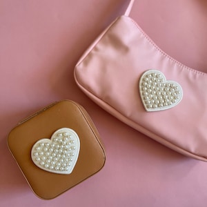 Puede incluir: Un bolso de mano rosa y un joyero marrón, ambos adornados con un parche blanco en forma de corazón adornado con pequeñas cuentas redondas con forma de perla. Los artículos están sobre un fondo rosa.