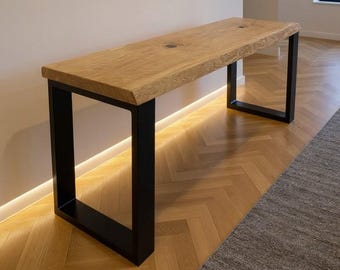 May include: A rectangular console table featuring a thick, natural-edge light brown wood slab top with visible grain and two dark circular inlays. The tabletop is supported by two sturdy, matte black metal U-shaped legs at each end.