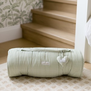 May include: A sage green and white gingham quilted roll featuring a horizontal zipper closure with a small heart-shaped fabric pull. A rectangular white label is centered on the front of the quilted fabric roll, which is resting on a textured rug.