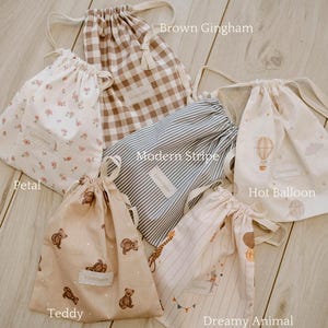 May include: A collection of drawstring bags in various patterns and colors. Designs include brown gingham, floral, blue and white stripes, hot air balloons, teddy bears, and a dreamy animal print. Each bag has a drawstring closure and a small label.