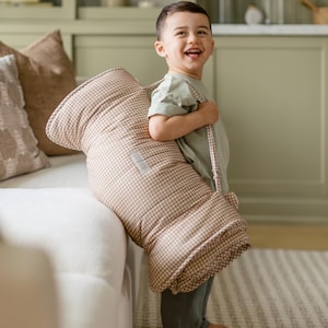 May include: A beige and white gingham patterned quilted nap mat with integrated carrying straps. The rectangular mat features a soft, padded texture and a small rectangular label on the front surface, designed for portability and comfort.