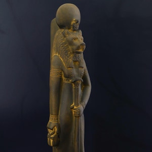 Könnte beinhalten: Eine dunkelbraune Statue der ägyptischen Göttin Sekhmet, dargestellt als löwenköpfige Frau. Sie hält einen Stab in ihrer rechten Hand und ein Symbol in ihrer linken Hand. Die Statue steht auf einem Sockel mit Hieroglyphen.