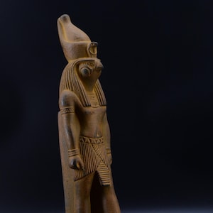 Może przedstawiać: Szczegółowa, zwietrzała statua egipskiego bóstwa, prawdopodobnie Horusa, z głową sokoła i wyszukanym nakryciem głowy. Postać stoi, z misteriowimi rzeźbami na podstawie i ubraniu. Statua ma ciepły, ziemisty brązowy kolor.
