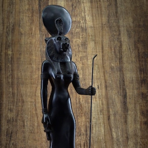 Może przedstawiać: Czarna drewniana statuetka egipskiej bogini Bastet, przedstawiona jako kobieta z głową lwa, nosząca dysk słoneczny i kobrę na głowie. Trzyma laskę w prawej ręce i symbol w lewej ręce. Statuetka stoi na podstawie z hieroglifami.