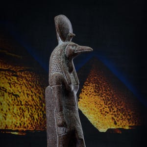 Könnte beinhalten: Eine dunkelbraune Steinstatue des ägyptischen Gottes Horus, dargestellt als falkenköpfiger Mann. Die Statue steht auf einem Sockel mit Hieroglyphen.