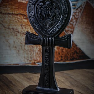 May include: A black Ankh statue with intricate carvings, including hieroglyphs and a central design. The Ankh is a symbol of life and is displayed on a rectangular base. The background features a patterned fabric.