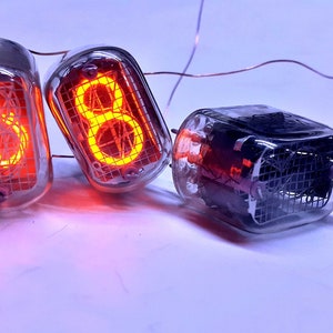 Może przedstawiać: Cztery zabytkowe lampy Nixie, dwie wyświetlające cyfry "6" i "8" w jasnym pomarańczowym kolorze, pozostałe dwie są wyłączone. Lampy są wykonane z przezroczystego szkła z metalową siatką wewnątrz i połączone miedzianym drutem.