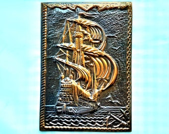 Può includere: Una placca da parete in metallo dorato che raffigura una nave a vela. La placca ha uno sfondo marrone scuro e un bordo a corda.