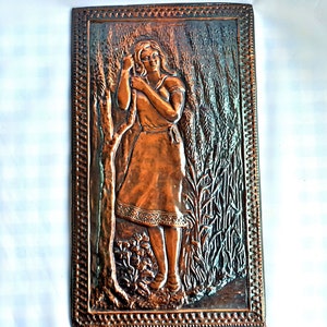 May include: A copper wall plaque with a raised relief design of a woman standing in a field of tall grass. The woman is wearing a long dress and has her hair pulled back. The plaque is framed with a decorative border.