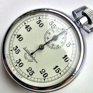 Puede incluir: Un cronómetro vintage con esfera blanca y números negros. La caja es de metal cromado y tiene una corona de cuerda. La esfera muestra el texto "Златоустовский Часовой Завод" y "СДЕЛАНО В СССР".