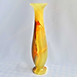 Pu&ograve; includere: Un vaso alto e sottile in onice giallo, arancione e marrone. Il vaso ha un'ampia apertura e una base stretta.