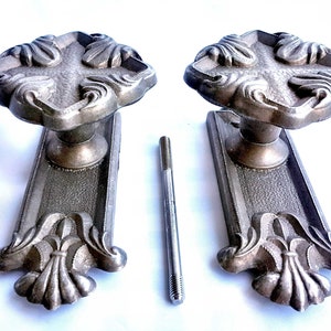 Pode incluir: Duas maçanetas de porta antigas com botões em forma de cruz ornamentados e placas decorativas. As maçanetas são de metal e têm um acabamento prateado. As maçanetas são fixadas a uma superfície branca com parafusos.