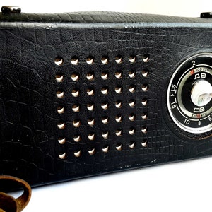 May include: A black leather vintage transistor radio with a silver dial and a brown leather strap. The radio has a speaker grille with a pattern of small holes.