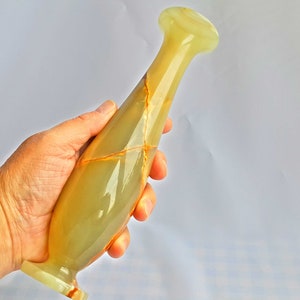 Pu&ograve; includere: Un vaso in onice verde e marrone con una forma slanciata e allungata. Il vaso ha una finitura liscia e lucida e un design delicato ed elegante.