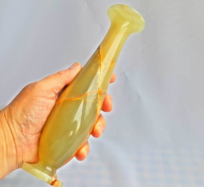 Pu&ograve; includere: Un vaso in onice verde e marrone con una forma slanciata e allungata. Il vaso ha una finitura liscia e lucida e un design delicato ed elegante.