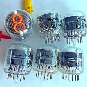 Peut inclure: Six tubes à vide en verre transparent avec des bases en métal noir et des fils rouges. Les tubes ont une grille et le texte "ИН-4" est visible sur le côté de chaque tube. Un tube est allumé avec une lueur orange, affichant le chiffre "8".