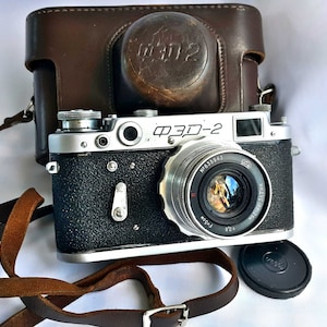 Puede incluir: Una cámara de película de 35 mm negra y plateada con una funda de cuero. La cámara tiene un objetivo con el texto "ФЭД-2" e "Industar-26" en él. La cámara está en buen estado y parece ser vintage.