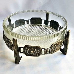 May include: A clear glass bowl with a decorative, patterned base. The bowl is on a metal stand with ornate details. The stand has three legs and a decorative band around the bowl.