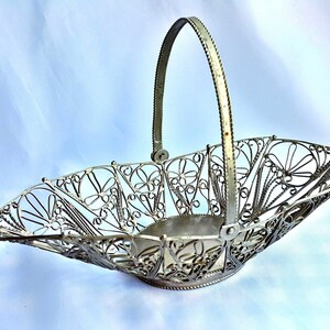 May include: A silver wire basket with a decorative floral design and a handle. The basket is empty and has a shiny finish.
