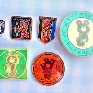 May include: A collection of seven vintage pins featuring the 1980 Moscow Olympics mascot, a brown bear named Misha. The pins are in various shapes and sizes, with different designs and colors. Some pins feature the bear with a bow and arrow, while others show the bear with a flag or standing alone. The pins are all in good condition and would be a great addition to any collection.