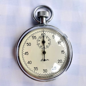 Può includere: Un orologio da tasca argentato con un quadrante bianco e lancette nere. L'orologio ha una lancetta dei secondi nera e un piccolo quadrante con numeri neri. Il quadrante dell'orologio ha segni neri per secondi e minuti. Il quadrante dell'orologio ha anche il testo "Am" e "СДЕЛАНО В СССР".