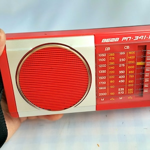 May include: A red and silver portable radio with a black strap. The radio has a dial with frequency markings in kHz and MHz. The radio is labeled "BEZA PN-391-1".