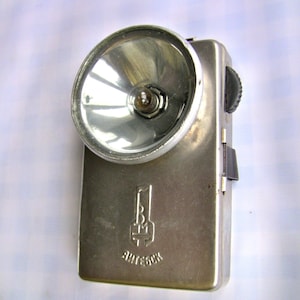 Könnte beinhalten: Eine silberne Taschenlampe mit einem runden Reflektor und einem schwarzen Schalter. Die Taschenlampe hat ein Logo mit der Aufschrift "BRETON" auf der Vorderseite.