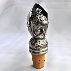 Puede incluir: Tapón de botella de plata con forma de cabeza de caballero que lleva un casco con detalles intrincados. El casco tiene una parte superior con bisagras y el caballero tiene bigote.