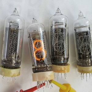Peut inclure: Quatre tubes Nixie en verre transparent avec des chiffres orange incandescents. Les tubes sont disposés en rangée et contiennent une grille métallique à l'intérieur. Les tubes sont montés sur une surface blanche.