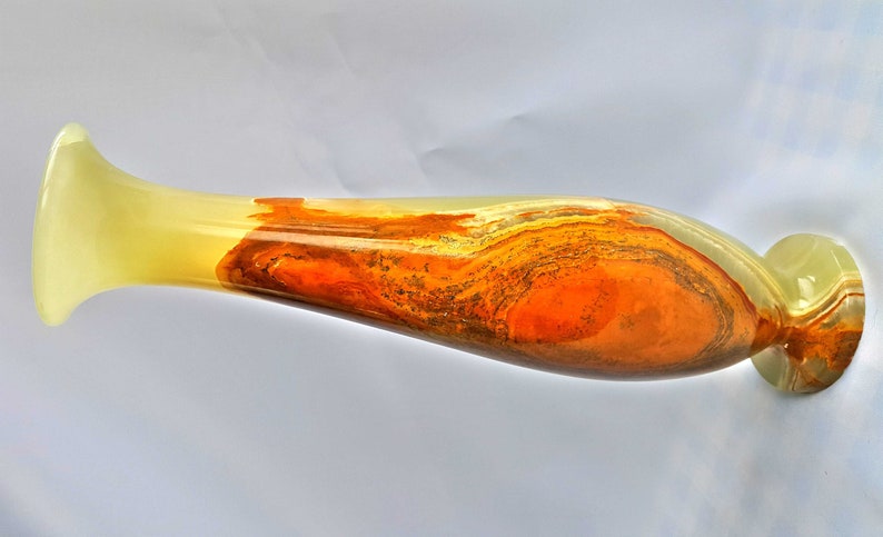 Pu&ograve; includere: Un vaso lungo e sottile in onice con una base giallo chiaro e un motivo a spirale di arancione, marrone e grigio.