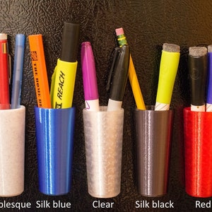 May include: A selection of pen holders in different colours, each containing writing implements. The holders are labelled "Marblesque", "Silk blue", "Clear", "Silk black", and "Red". The image displays pens, pencils, and markers.