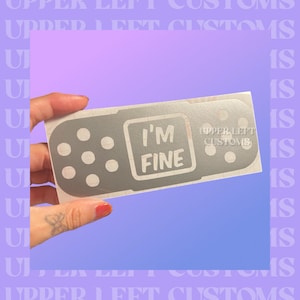 May include: A silver bandage-shaped sticker with the words "I'M FINE" in a square. The sticker has several small, evenly spaced circles and the text "UPPER LEFT CUSTOMS" on the right side. The sticker is held against a purple and blue gradient background.