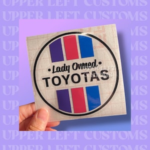 May include: A round sticker with a black outline features the text "Lady Owned TOYOTAS." The design includes vertical stripes in purple, red, and blue. The sticker is held against a purple background.