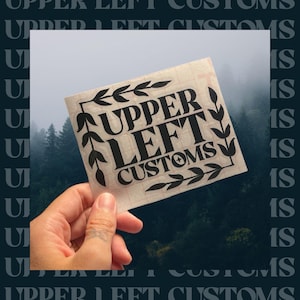 May include: A square sticker with the text "UPPER LEFT CUSTOMS" in a bold, black font, framed by decorative leaf and arrow designs. The sticker is held against a backdrop of a misty forest.