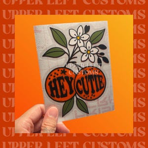 May include: A sticker featuring two orange-colored oranges with the text "HEY CUTIE" in black. The design includes white flowers, green leaves, and the text "UPPER LEFT CUSTOMS". The sticker is held against an orange background.