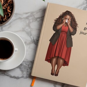 K&ouml;nnte beinhalten: Ein beigefarbenes Notizbuch mit einer gedruckten Illustration einer Person mit langem dunklem Haar, die ein rotes Kleid und einen dunklen Blazer tr&auml;gt und ein Taschentuch vor das Gesicht h&auml;lt. Der Text "my grief journey" steht in Schwarz auf dem Cover.