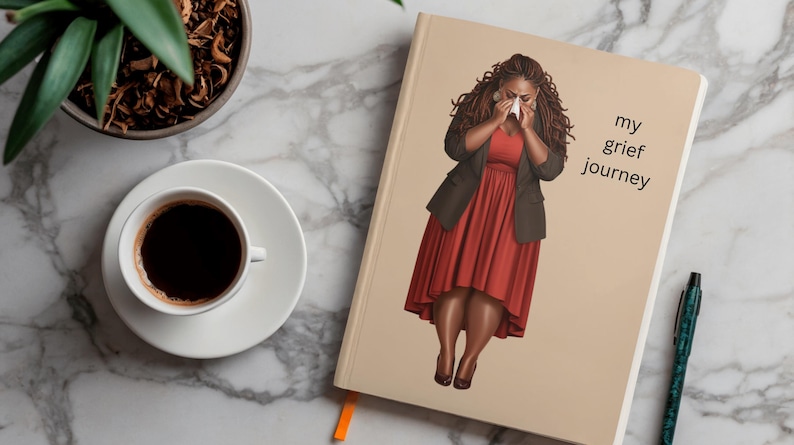 K&ouml;nnte beinhalten: Ein beigefarbenes Notizbuch mit einer gedruckten Illustration einer Person mit langem dunklem Haar, die ein rotes Kleid und einen dunklen Blazer tr&auml;gt und ein Taschentuch vor das Gesicht h&auml;lt. Der Text "my grief journey" steht in Schwarz auf dem Cover.
