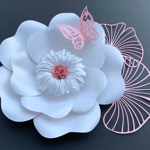 May include: A large white paper flower with a pink center and a pink butterfly decoration. The flower is made of multiple layers of paper and has a delicate, layered look. The butterfly is cut out of paper and has a delicate, intricate design.