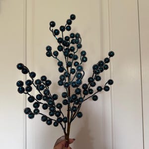 May include: A decorative floral spray featuring numerous small, round berries in a deep teal blue color. Each berry is coated in a fine, sparkling glitter texture. The berries are attached to thin, brown, flexible wire stems that branch out.