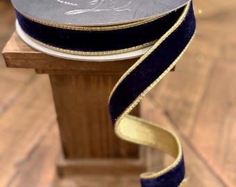 May include: A spool of dark navy blue velvet ribbon featuring metallic gold edges. The ribbon is wound around a circular gray spool with a white label and a silver logo. A portion of the ribbon hangs down, displaying the deep blue velvet texture and the gold trim on both sides.