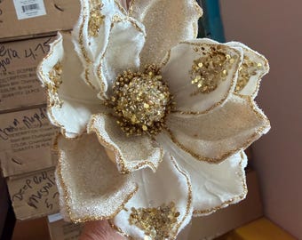 May include: A decorative artificial flower featuring large, cream-colored petals with gold glitter-dusted edges. The center of the flower is densely packed with small, sparkling gold beads and sequins, creating a textured, shimmering appearance.