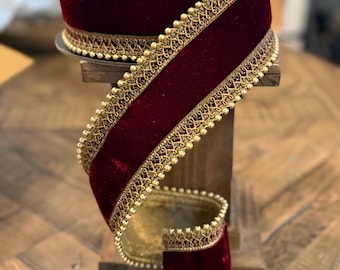 Könnte beinhalten: Eine Rolle tiefrotes Samtband mit aufwendiger goldener Metallverzierung an beiden Kanten. Das Band ist mit einer feinen Reihe kleiner goldener Perlen entlang der Ränder verziert, was für ein strukturiertes und verziertes Erscheinungsbild sorgt.