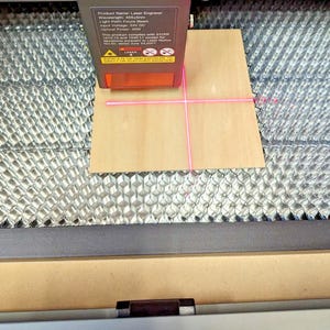 May include: A laser engraver with a wooden plank in place. The laser beam emits a pink crosshair onto the wood. The engraver has a silver and black metal casing. The product name is visible on the engraver.