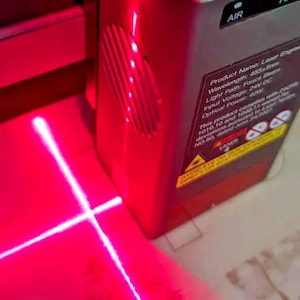 May include: A red laser level projects a bright red crosshair onto a surface. The device has a red casing and a label with technical specifications. The laser beam is clearly visible, indicating its alignment function.