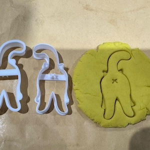 Può includere: Due stampini per biscotti in plastica bianca a forma di gatto con la coda in aria, e un ritaglio di pasta per biscotti gialla della stessa forma di gatto.