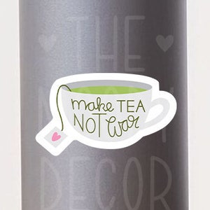 May include: White teacup with green tea inside and a pink heart-shaped tea bag. The teacup has the text "Make tea not war" written on it.